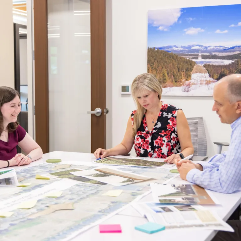 Three colleagues in a modern conference room review maps and documents covered with sticky notes, discussing plans around a white table beneath a wall photo of a snowy mountain landscape.