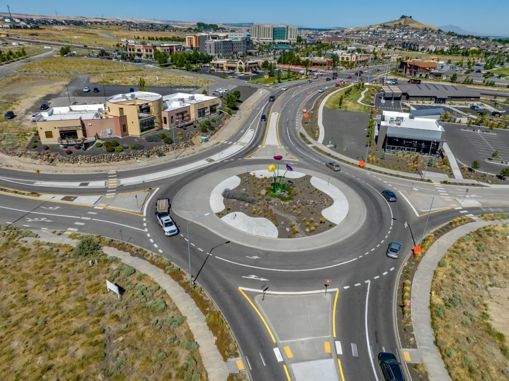Aerial view of a multilane roundabout in a suburban commercial area, with cars circulating and a central island landscaped with tall colorful flower sculptures.