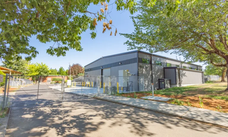 Gray metal warehouse behind a chain-link fence and gated entrance, with roll-up doors and yellow bollards, along a tree-lined street on a sunny day.