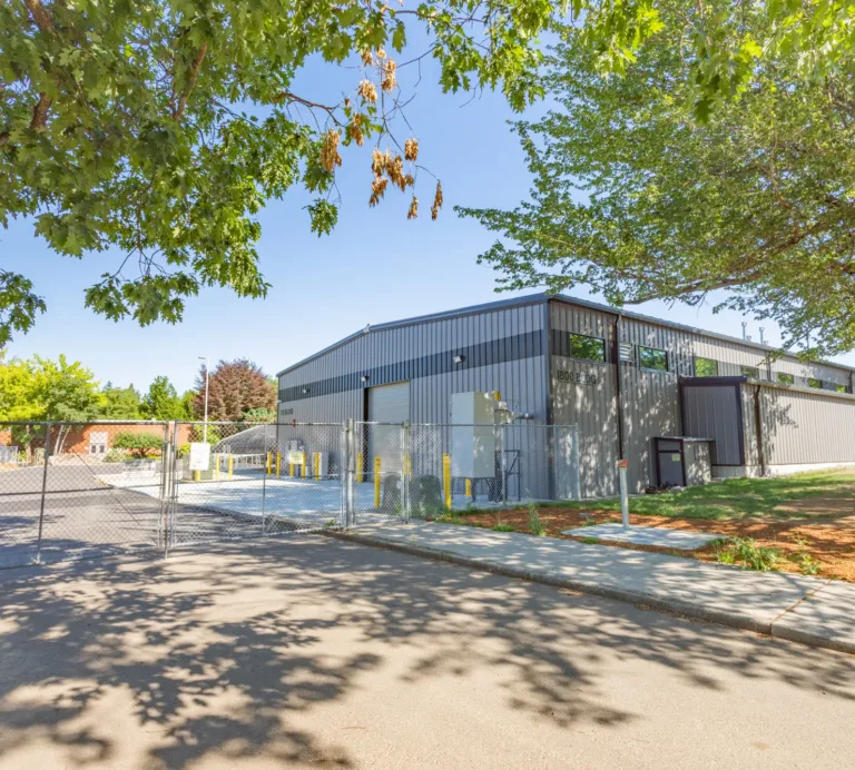 Gray metal warehouse behind a chain-link fence and gated entrance, with roll-up doors and yellow bollards, along a tree-lined street on a sunny day.