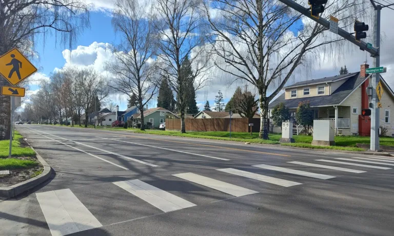 Wide residential street intersection with a freshly painted zebra crosswalk, yellow pedestrian-crossing signs and overhead signals, leafless trees lining the road, and houses under a blue sky with clouds; the corner street sign reads “Washington Way.”
