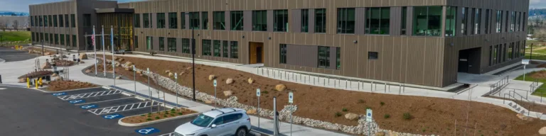 A modern two-story office building with large dark-framed windows and a flat roof, fronted by a parking lot with multiple accessible spaces, a single parked SUV, flagpoles by the entrance, and newly landscaped grounds under a partly cloudy blue sky.