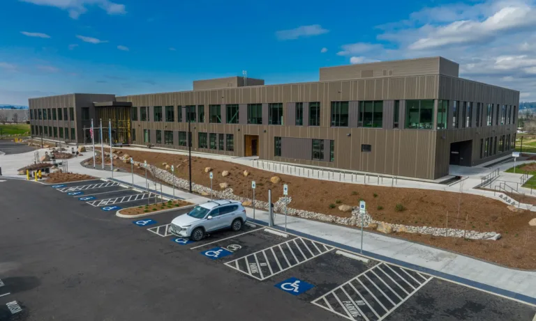 A modern two-story office building with large dark-framed windows and a flat roof, fronted by a parking lot with multiple accessible spaces, a single parked SUV, flagpoles by the entrance, and newly landscaped grounds under a partly cloudy blue sky.