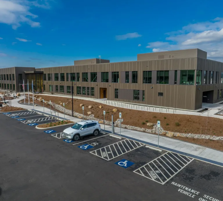A modern two-story office building with large dark-framed windows and a flat roof, fronted by a parking lot with multiple accessible spaces, a single parked SUV, flagpoles by the entrance, and newly landscaped grounds under a partly cloudy blue sky.