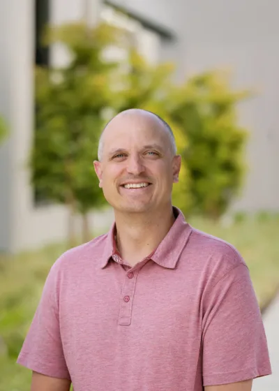 Headshot of Jason Irving smiling outdoors in a pink polo, with a shaved head and a blurred leafy background.