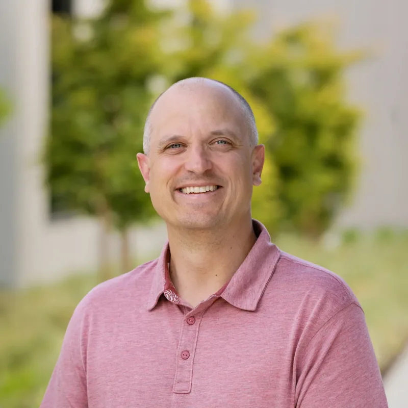 Headshot of Jason Irving smiling outdoors in a pink polo, with a shaved head and a blurred leafy background.