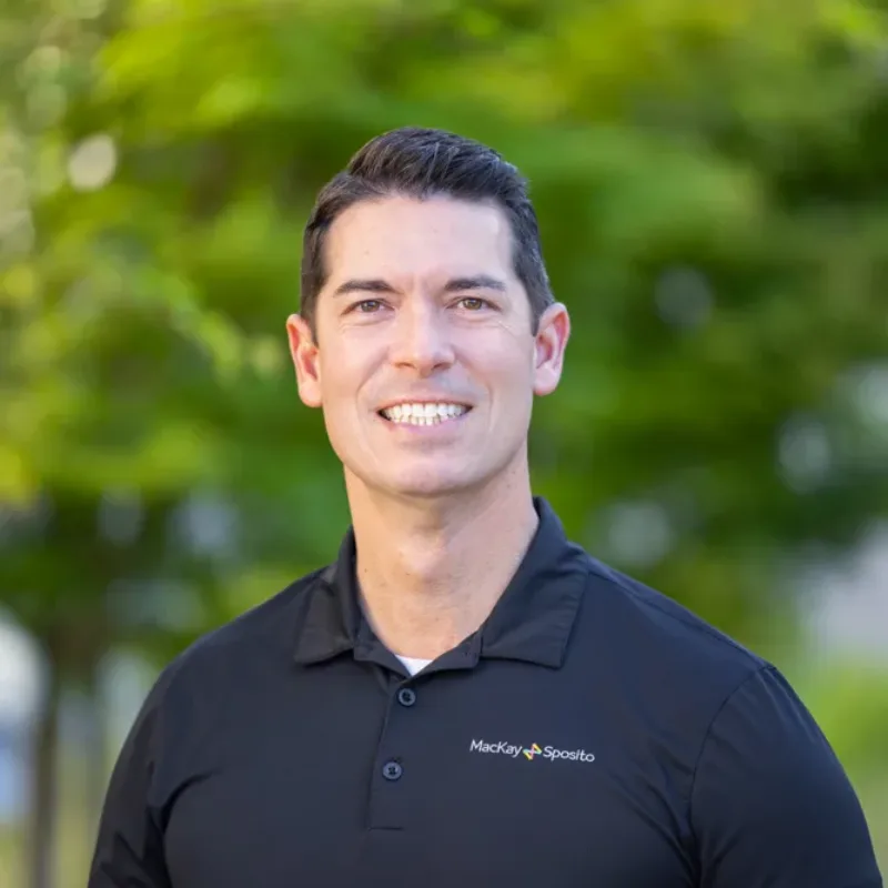 Headshot of Jason Joyce smiling in a black polo with the MacKay Sposito logo, outdoors against a blurred green background.