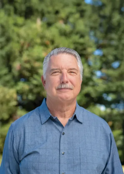 Headshot of Jerry Aplass outdoors, with short gray hair and a mustache, wearing a light blue button-down shirt against a blurred background of green trees.