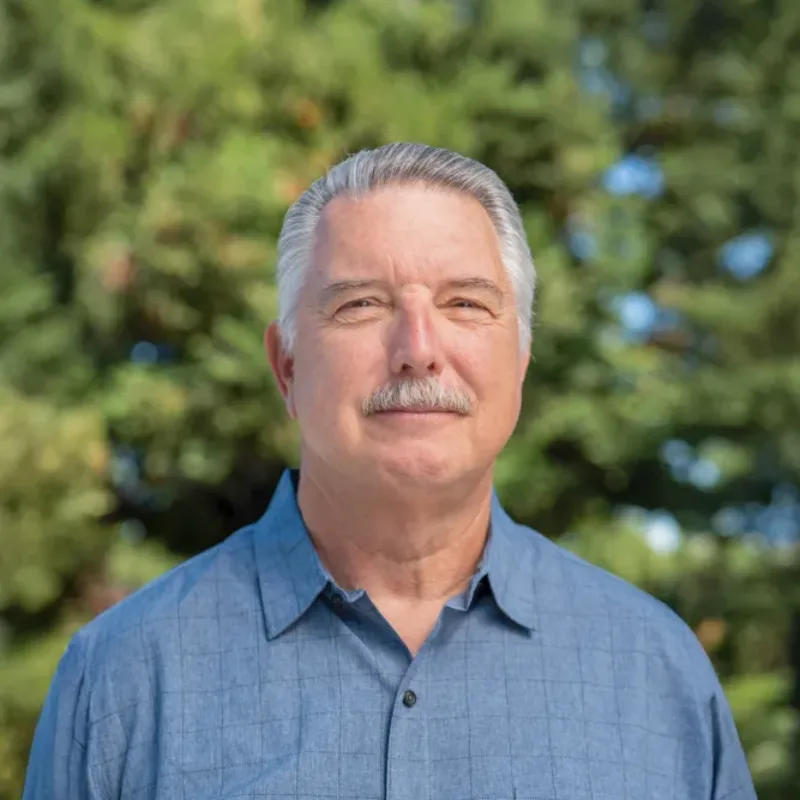Headshot of Jerry Aplass outdoors, with short gray hair and a mustache, wearing a light blue button-down shirt against a blurred background of green trees.
