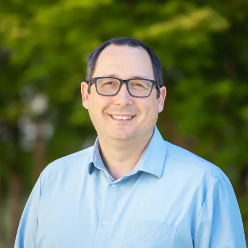 Headshot of Jerry Balch, a smiling person with short dark hair and glasses wearing a light blue button-down shirt against a blurred green foliage background.