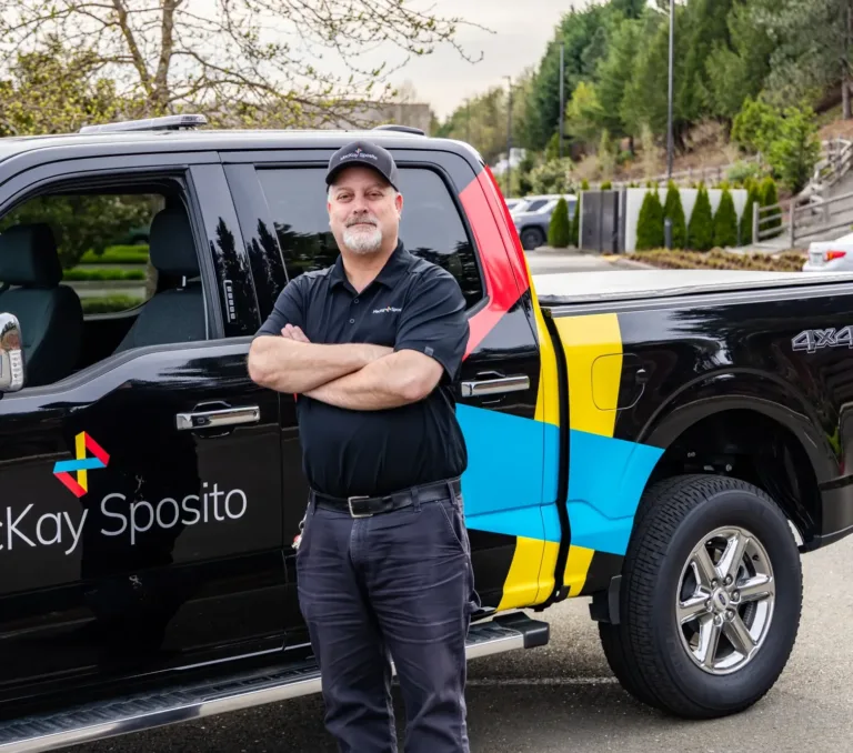 MacKay Sposito team member with arms crossed stands beside a branded black pickup truck with colorful graphics in an outdoor parking area.