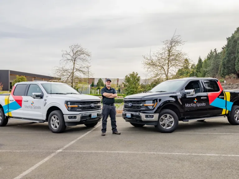 Two MacKay Sposito–branded Ford pickup trucks with an employee standing between them in a parking lot, with trees and office buildings in the background.