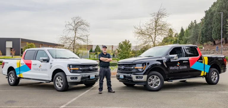 Person standing between a white and a black Ford pickup, both wrapped with MacKay Sposito branding and colorful geometric graphics, parked outdoors at a business campus.