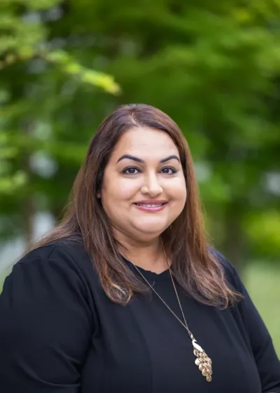 Headshot of Jyoti Sohi, smiling outdoors in a black top with a gold pendant necklace against a blurred green background.