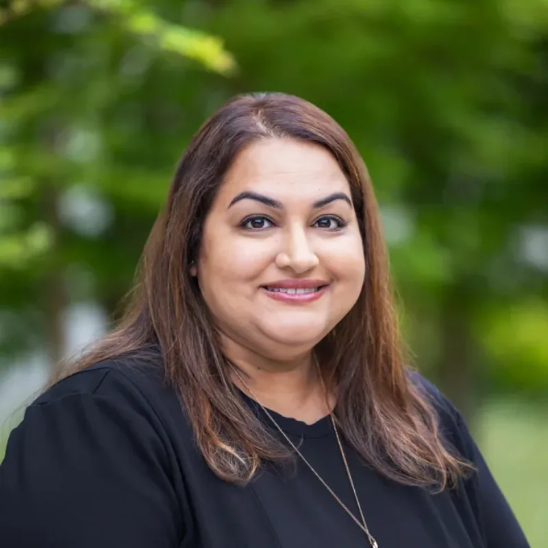 Headshot of Jyoti Sohi, smiling outdoors in a black top with a gold pendant necklace against a blurred green background.