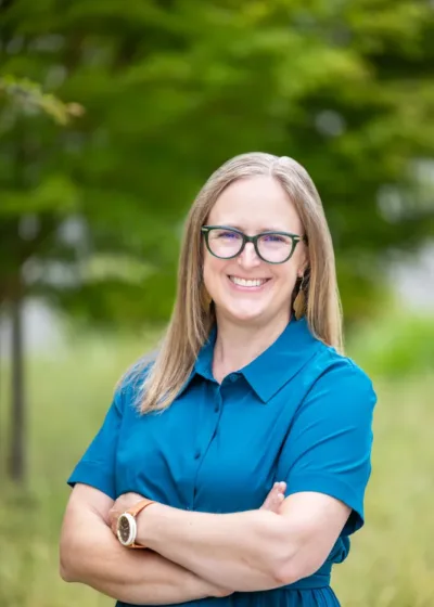 Headshot of Kristen Currens smiling with glasses and shoulder-length hair, wearing a teal dress with arms crossed against a blurred green foliage background.