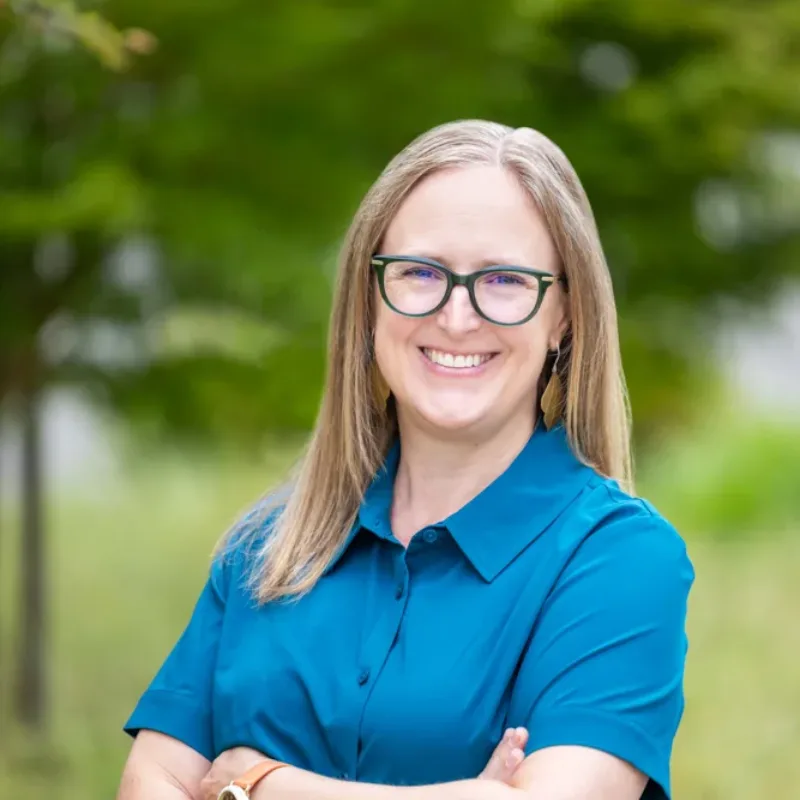 Headshot of Kristen Currens smiling with glasses and shoulder-length hair, wearing a teal dress with arms crossed against a blurred green foliage background.
