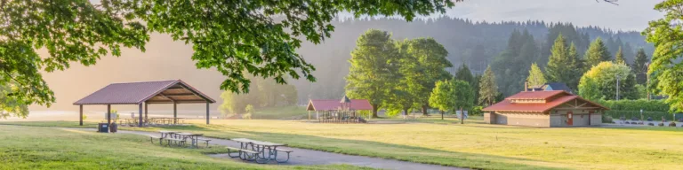 Early morning light over a grassy park with a covered picnic shelter, picnic tables along a paved path, a playground and restroom building, framed by leafy branches and forested hills in the background.