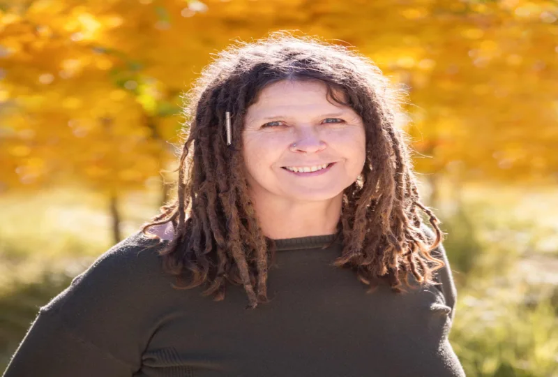 Headshot of Lee Lynn Michel, a smiling person with long brown dreadlocks and a small nose ring, wearing a dark green sweater with golden autumn foliage softly blurred in the background.