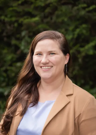 Headshot of Lisa Moore smiling outdoors, with long brown hair, wearing a tan blazer over a light blue top against a leafy green background.
