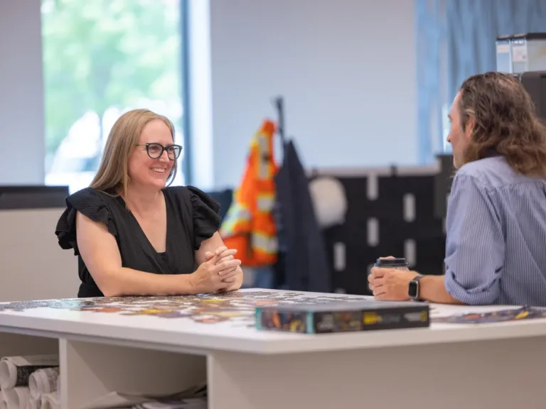 Two MacKay Sposito colleagues talk at an office table; one holds a coffee cup, with a high‑visibility vest and hard hat blurred in the background.