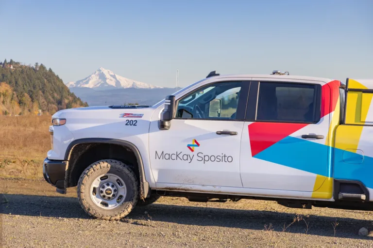 MacKay Sposito–branded white pickup truck with colorful geometric graphics parked on a gravel road, with a snow-capped mountain and clear blue sky in the background.