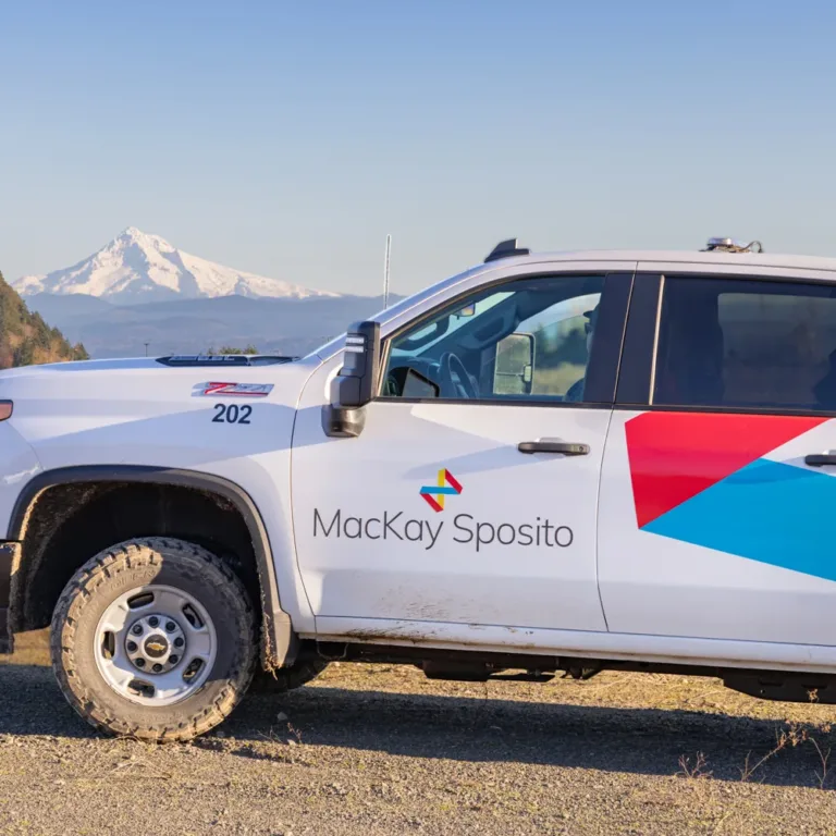MacKay Sposito-branded pickup truck with colorful geometric graphics parked on gravel, with a snow-capped mountain and forested hills under a clear blue sky in the background.