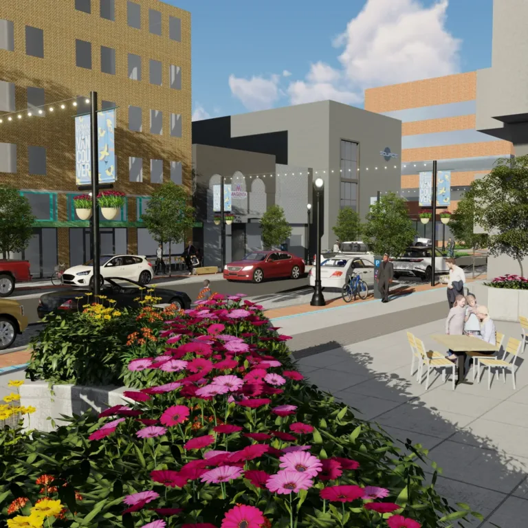 Rendering of a downtown street with bright flower planters, outdoor café seating, string lights, cars and bikes, and “Vancouver” banners among modern buildings on a sunny day.