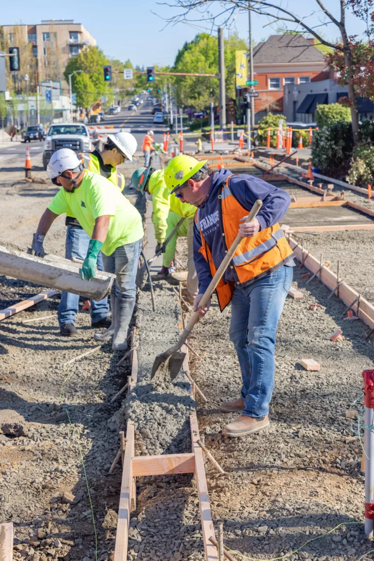 Construction crew in hard hats and safety vests pour and spread concrete into wooden forms for a sidewalk along a city street lined with traffic cones.