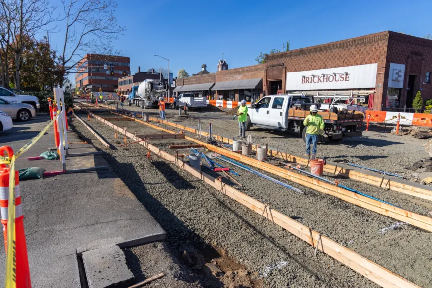Construction workers repair a downtown street, setting wooden forms and rebar for new curbs as trucks and a concrete mixer operate beside orange safety barriers and storefronts under a clear sky.