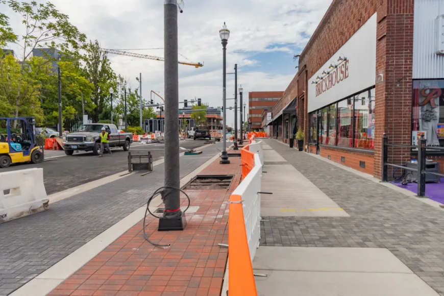 City street undergoing construction with orange-and-white barriers, workers and trucks, new lampposts and brick pavers, and a row of storefronts including one labeled “BRICKHOUSE.”