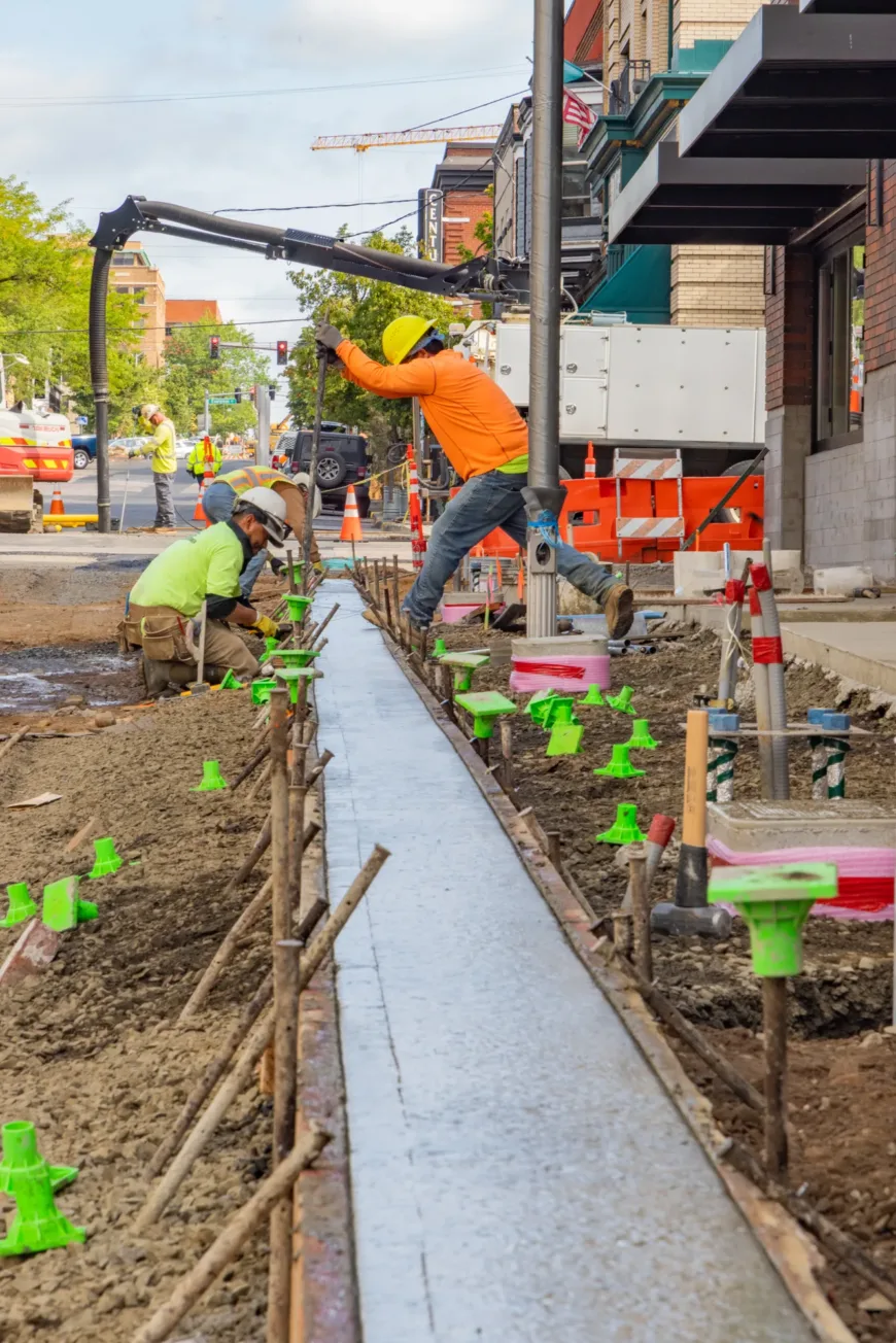 Construction workers in hard hats pour and level wet concrete into a narrow sidewalk form using a pump hose along a city street lined with traffic cones and equipment.