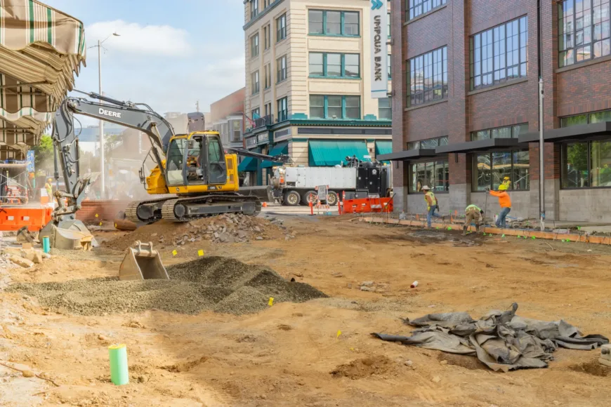 Downtown street construction scene with a yellow excavator and crew forming a sidewalk beside brick buildings, surrounded by orange barriers, cones, and piles of dirt and gravel.