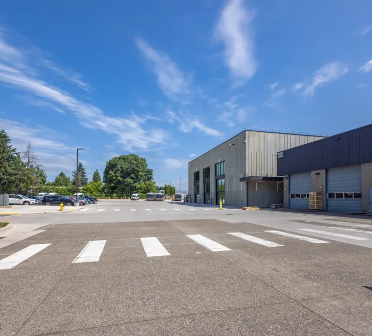 Industrial warehouse exterior with multiple roll-up bay doors and a 'Shipping & Receiving' area, facing a wide paved lot with crosswalks, parked cars, and trees under a bright blue sky.