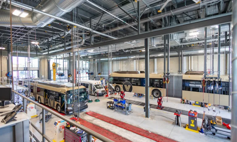 Large, modern bus maintenance facility with multiple tan city buses—one articulated—parked in service bays surrounded by lifts, hoses, and workshop equipment under high exposed ducts.