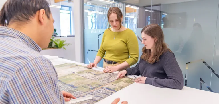 MacKay Sposito team reviewing a large aerial site plan together in a bright, modern office with glass walls.