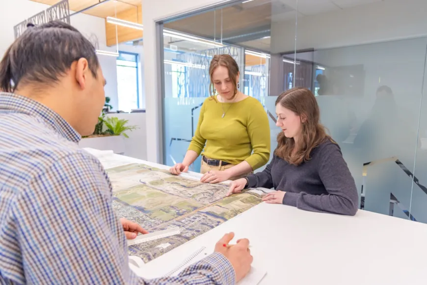MacKay Sposito team reviewing a large aerial site plan together in a bright, modern office with glass walls.