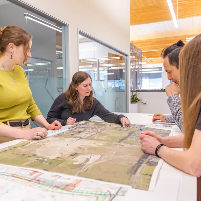 MacKay Sposito team collaborating around a conference table, reviewing large aerial site maps and plan drawings in a modern glass-walled office.