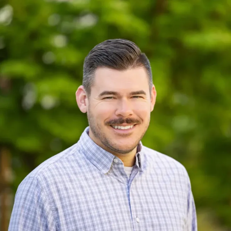 Headshot of Michael Strachan smiling, with short dark hair and a mustache, wearing a light plaid button-down shirt against a blurred green outdoor background.