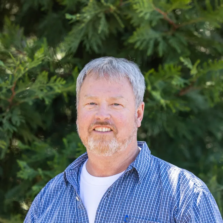Headshot of Monte Grondahl smiling outdoors, a person with short gray hair and a trimmed beard wearing a blue checkered shirt over a white T-shirt, with leafy evergreens in the background.