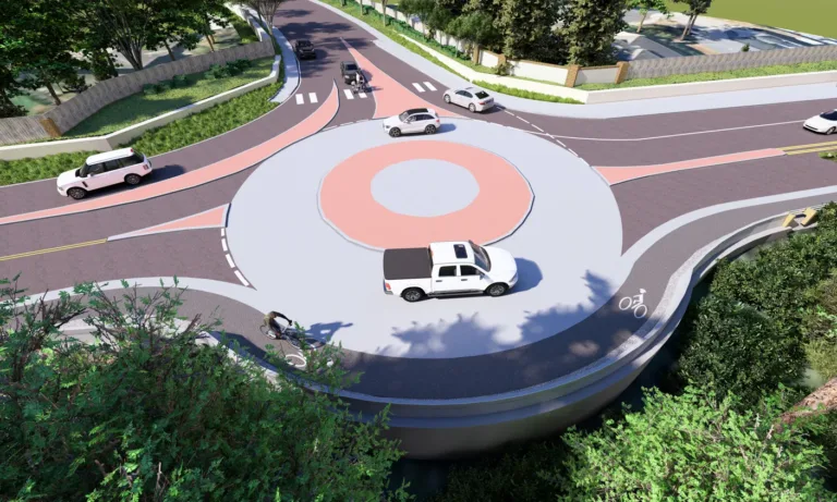 Aerial rendering of a landscaped roundabout with multiple cars circulating, a cyclist in an outer bike lane, and marked pedestrian crosswalks at each approach.