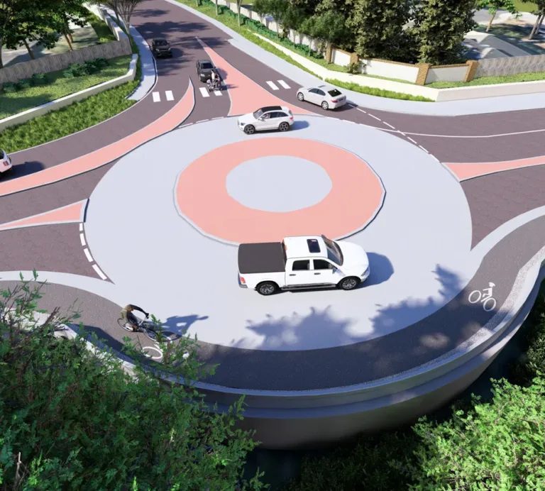 Aerial rendering of a landscaped roundabout with multiple cars circulating, a cyclist in an outer bike lane, and marked pedestrian crosswalks at each approach.
