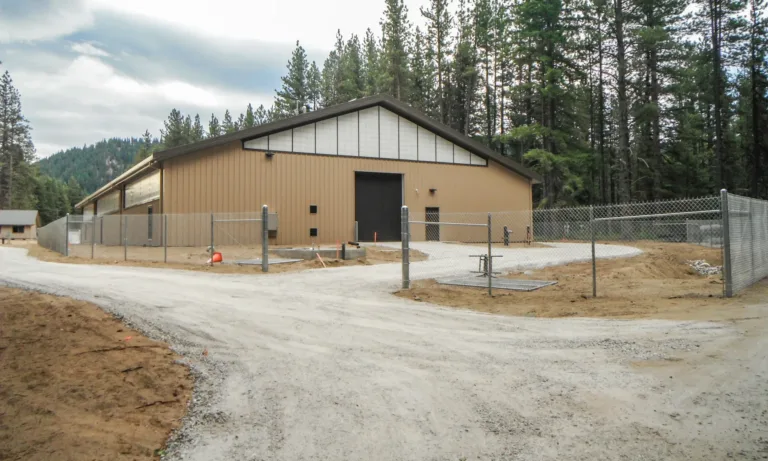 Large tan metal warehouse with a gabled roof and roll-up door, surrounded by chain-link fencing along a gravel road in a pine-forested, mountainous area under an overcast sky.