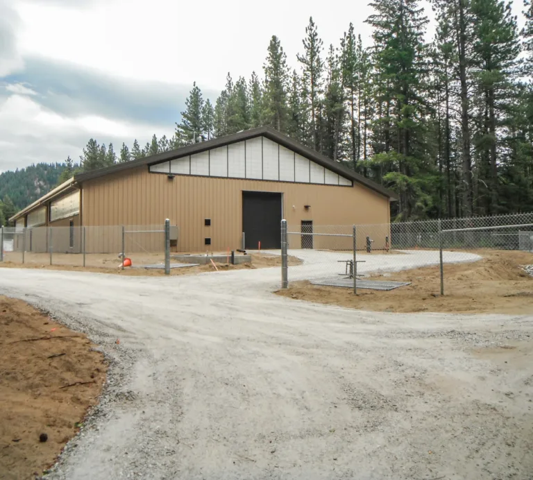 Large tan metal warehouse with a gabled roof and roll-up door, surrounded by chain-link fencing along a gravel road in a pine-forested, mountainous area under an overcast sky.