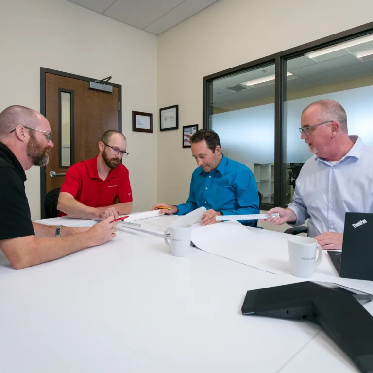 MacKay Sposito team members collaborate in a conference room, reviewing large engineering plans with drafting scales, mugs, and a laptop on the table.