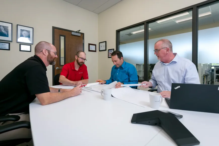 MacKay Sposito team members collaborate in a conference room, reviewing large engineering plans with drafting scales, mugs, and a laptop on the table.
