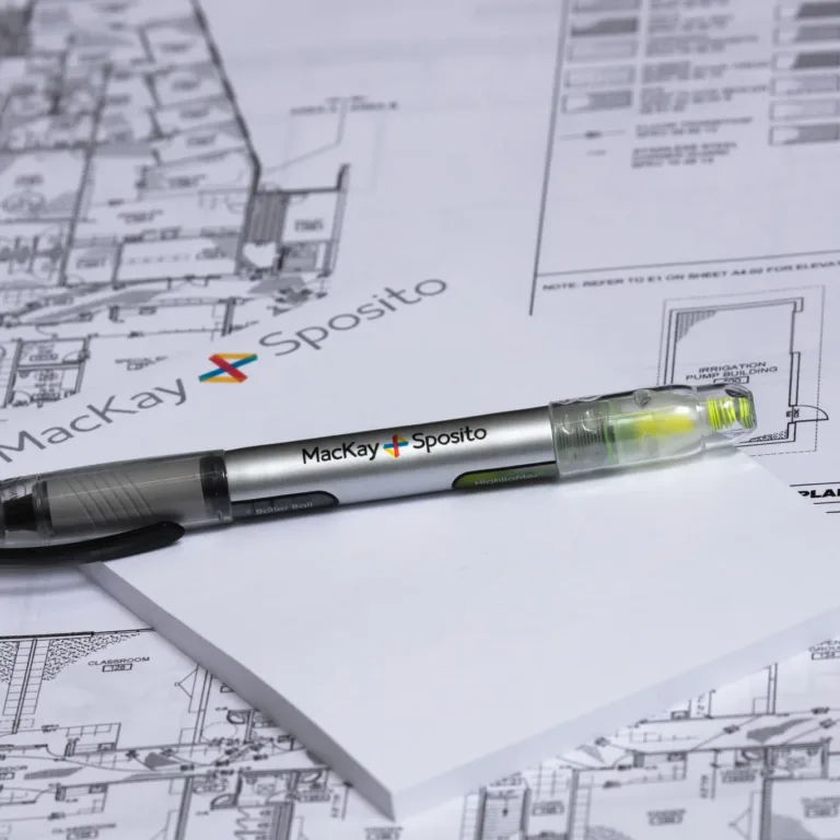 Historical photo of a MacKay Sposito–branded pen/highlighter on a notepad atop architectural blueprints, with the MacKay Sposito logo visible in the background.