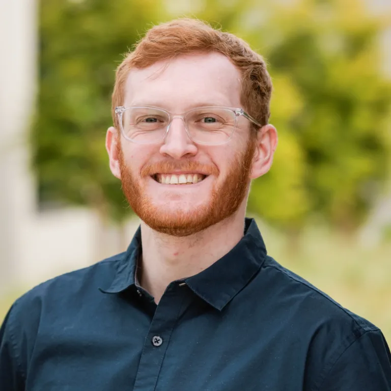 Headshot of Paton Malley smiling, with short red hair, a red beard, and clear-framed glasses, wearing a dark button-up shirt against a blurred green outdoor background.