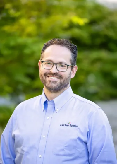 Headshot of Paul Smith, a smiling person with glasses and short dark hair, wearing a light blue MacKay Sposito button-down against a green, leafy background.