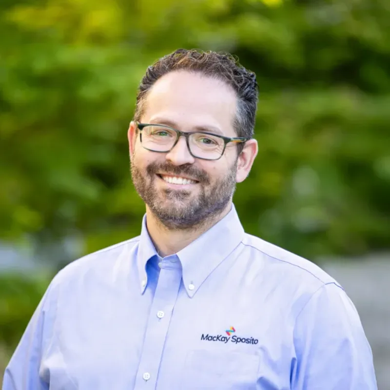 Headshot of Paul Smith, a smiling person with glasses and short dark hair, wearing a light blue MacKay Sposito button-down against a green, leafy background.